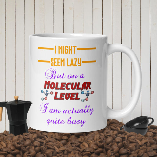 White glossy mug with humorous molecular design and text, surrounded by coffee beans and a small coffee maker, on a wooden background.