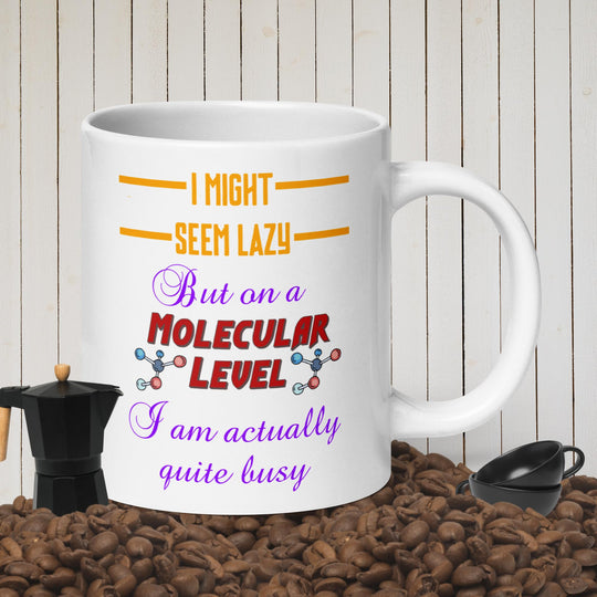 White glossy mug with colorful "I might seem lazy but on a molecular level I am actually quite busy" text, surrounded by coffee beans and brewing tools.